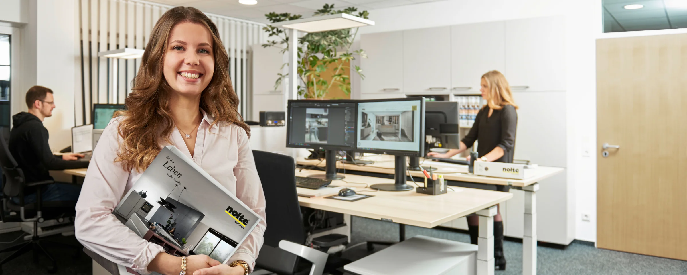 In un ufficio moderno, persone lavorano al computer mentre una donna tiene un catalogo.