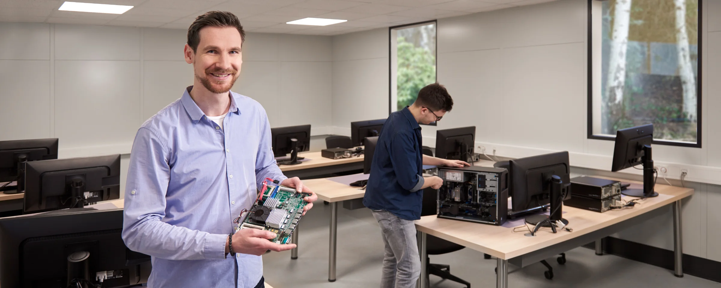 Un uomo tiene un componente del computer in un'aula con diversi computer e un altro uomo sullo sfondo.