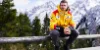 Un uomo con una giacca gialla seduto su una staccionata di legno davanti a montagne innevate.