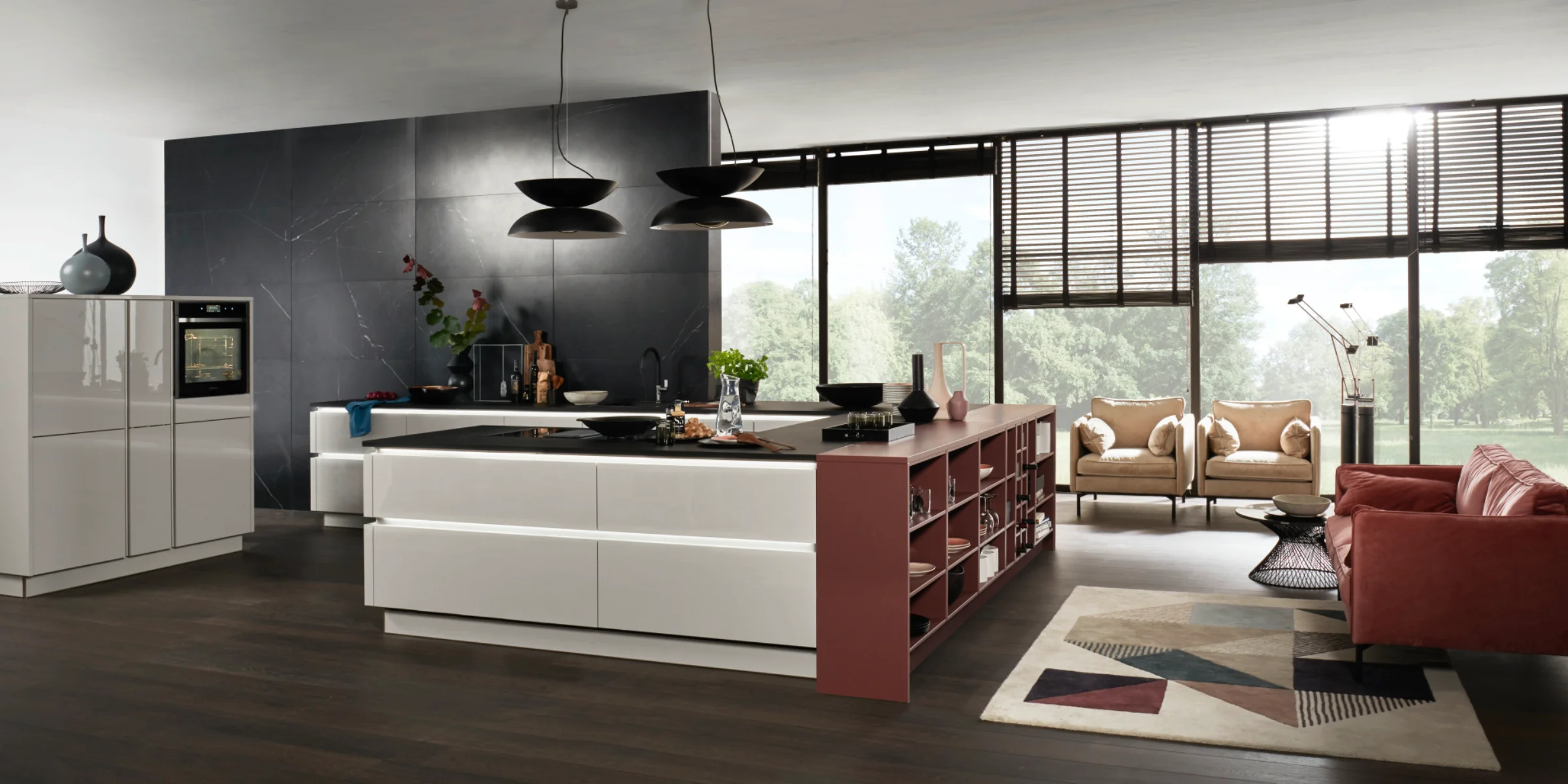 Moderna Nolte cucina con il frontale lucido NOVA LACK Platingrau. La cucina a isola senza maniglie colpisce per le linee chiare, un ampio piano di lavoro con piano cottura integrato e uno spazio aperto a scaffale in caldo rosso bruno. L'ambiente appare aperto e inondato di luce grazie a finestre a tutta altezza e un arredo elegante con un'accogliente area salotto in stile soggiorno.