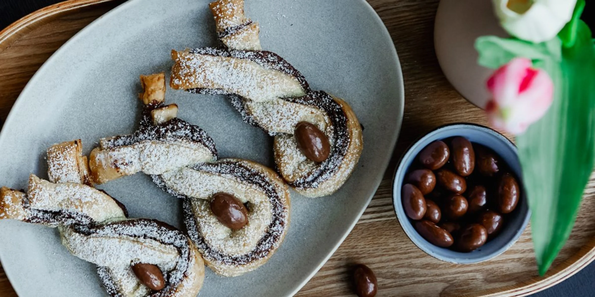 Tre dolci al cioccolato su un piatto con mandorle, accanto un vaso di fiori.