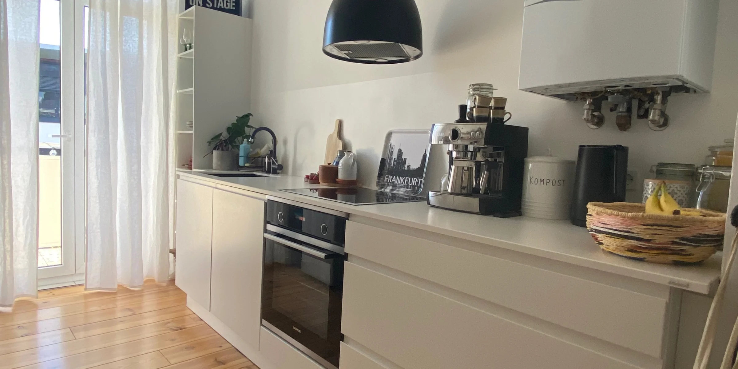 La moderna cucina Nolte con il programma SIGMA LACK si trova in una stanza luminosa con grandi finestre e tende bianche lunghe fino al pavimento. I mobili e cassetti bianchi senza maniglie si armonizzano con il caldo pavimento in legno. Un forno da incasso con piano cottura nero è posizionato al centro, affiancato da un lavello con rubinetteria scura. Sul piano di lavoro, integrato da parte del cliente, si trovano una macchina per espresso, una caraffa nera e un contenitore per compost. Una lampada sospesa nera crea un accento moderno. Dettagli personali come un'insegna „ON STAGE“ e un vassoio Frankfurt-Skyline completano l'ambiente.