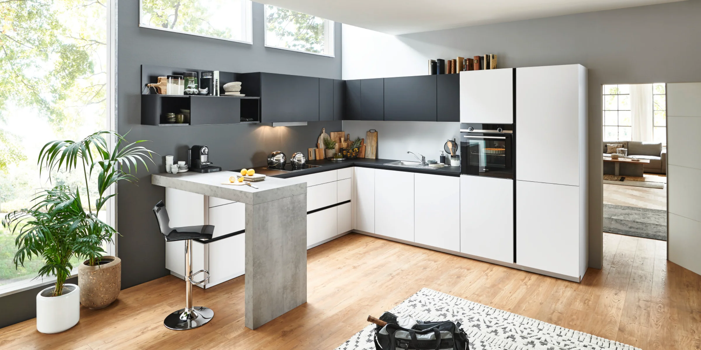 Moderna Nolte cucina in L-Form und banco con FEEL fronti in Arcticweiß softmatt e PLUS fronti in Schwarz softmatt. Il design senza maniglie è completato da un sorprendente bancone effetto cemento. L'effetto aperto e luminoso della stanza è sostenuto da molta luce naturale e materiali naturali come pavimento in legno e piante – uno spazio elegante e funzionale per cucinare e vivere.
