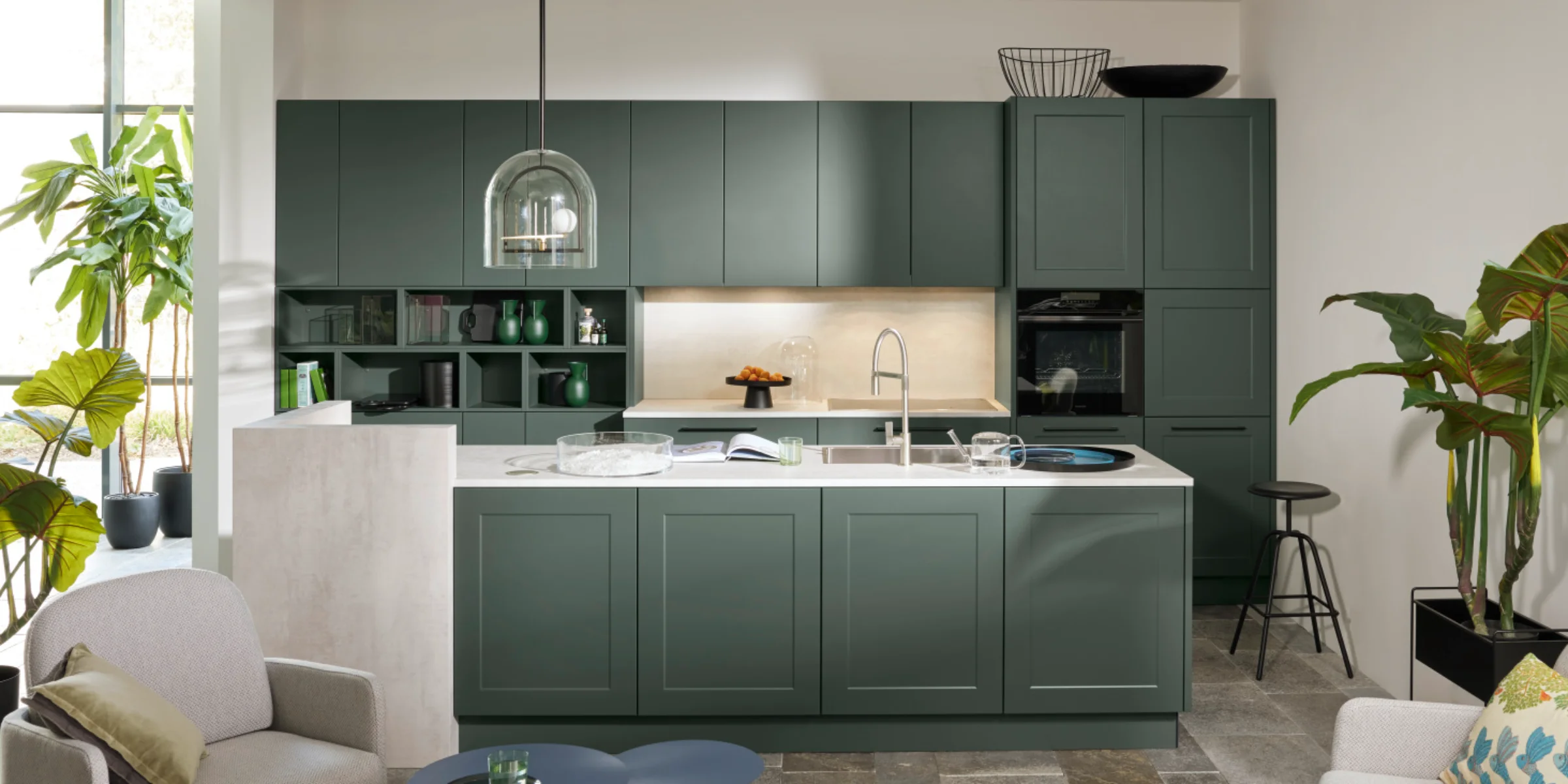 Moderna cucina Nolte con una combinazione di frontali FRAME LACK e SOFT LACK in Black Green softmatt, isola di cottura con design a cornice e piano di lavoro chiaro in Beton Weiß, combinata con accogliente area soggiorno.