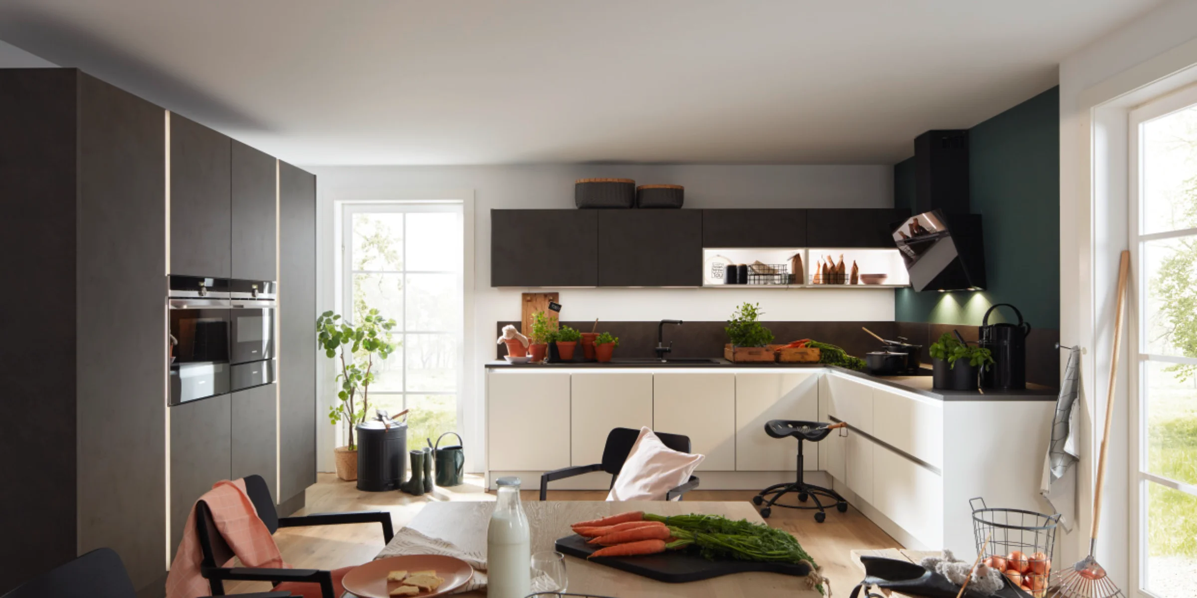 Moderna Nolte Küche con il fronte distintivo PORTLAND in Cemento Antracite e il fronte Antifingerprint PLUS in Bianco soft opaco. La cucina a due colori combina pensili scuri con armadi inferiori chiari e offre un design senza maniglie e facile da mantenere. Ampie finestre, dettagli in legno ed elementi decorativi naturali creano un'atmosfera accogliente e luminosa.