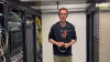 Un uomo in una sala server tra scaffali pieni di cavi e attrezzature tecnologiche.
