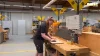 Una persona lavora a un tavolo di legno con attrezzi in un'officina; sullo sfondo si vedono altre postazioni di lavoro.