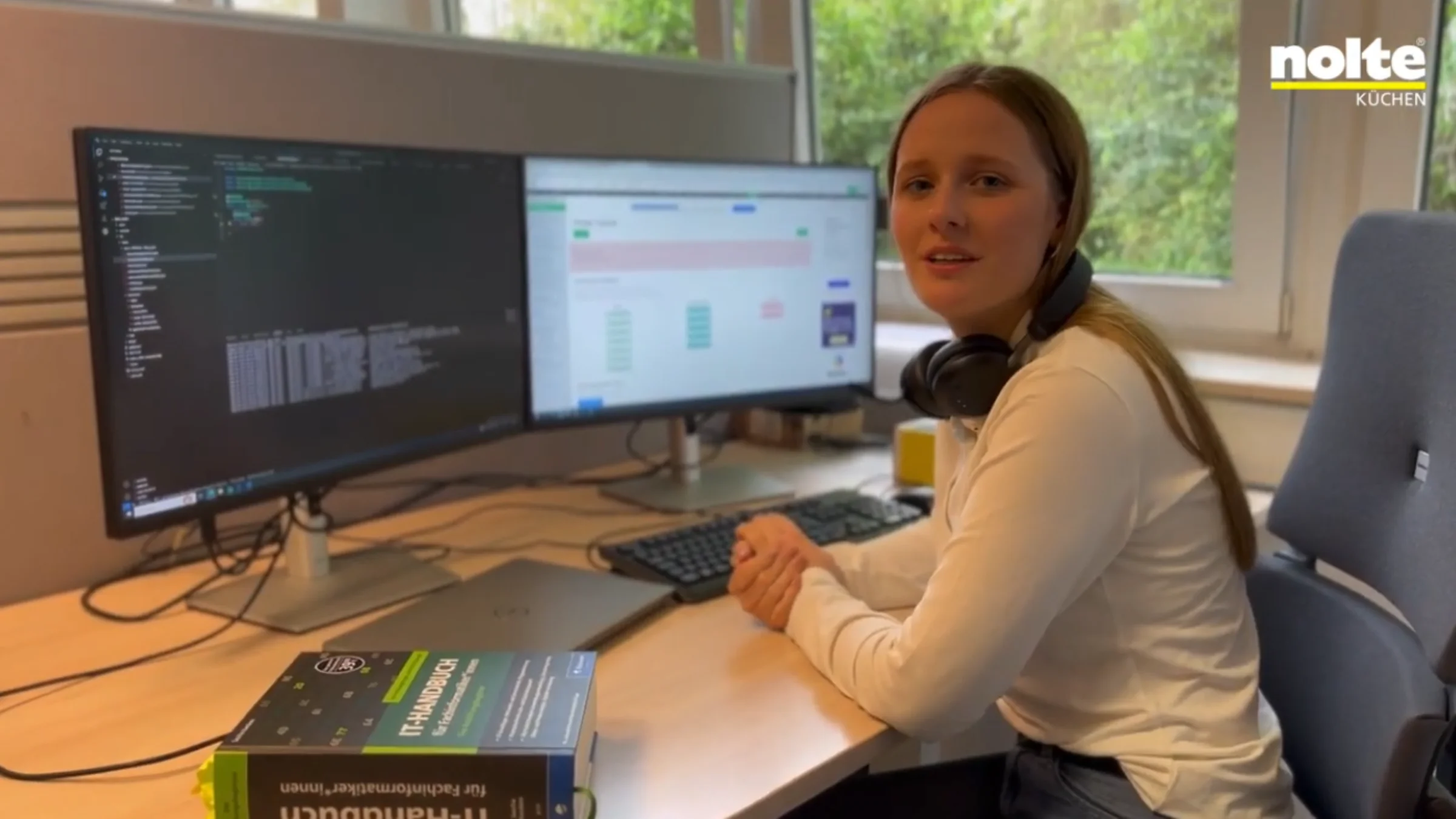 Una donna è seduta a una scrivania con due monitor in ufficio, a sinistra un libro sull'IT.