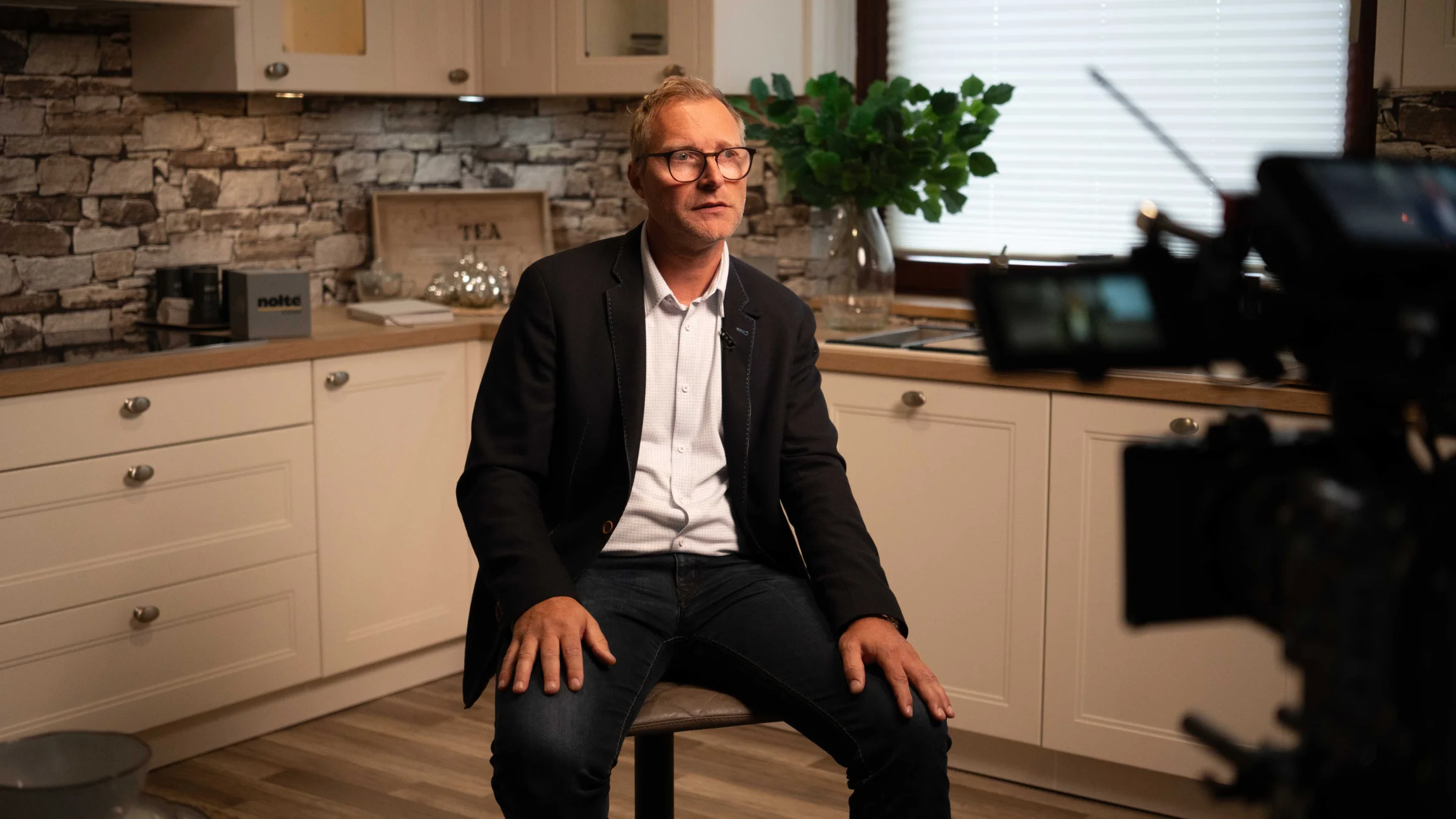 Un uomo siede in una cucina moderna con parete in pietra e armadi bianchi. Nolte Kitchen.