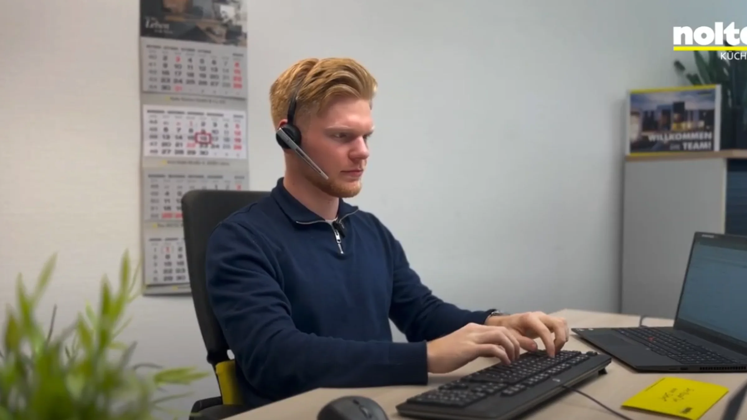 Un uomo lavora concentrato a una scrivania con computer e cuffie; sullo sfondo il logo di Nolte Kitchen.