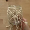 Eine Hand hält ein Glas mit Makramee-Dekor auf einem Holztisch.