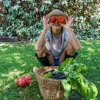 Persona tiene tomates frente a su cara, sentada en el jardín junto a una cesta de verduras.
