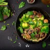 Piatto nero con insalata di quinoa, broccoli, funghi e foglie di basilico su un tavolo scuro.