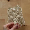 Una mano tiene un vaso con decoración de macramé sobre una mesa de madera.