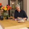 Un uomo siede al bancone della cucina con fiori e macarons, leggendo una rivista. Nolte Kitchen.
