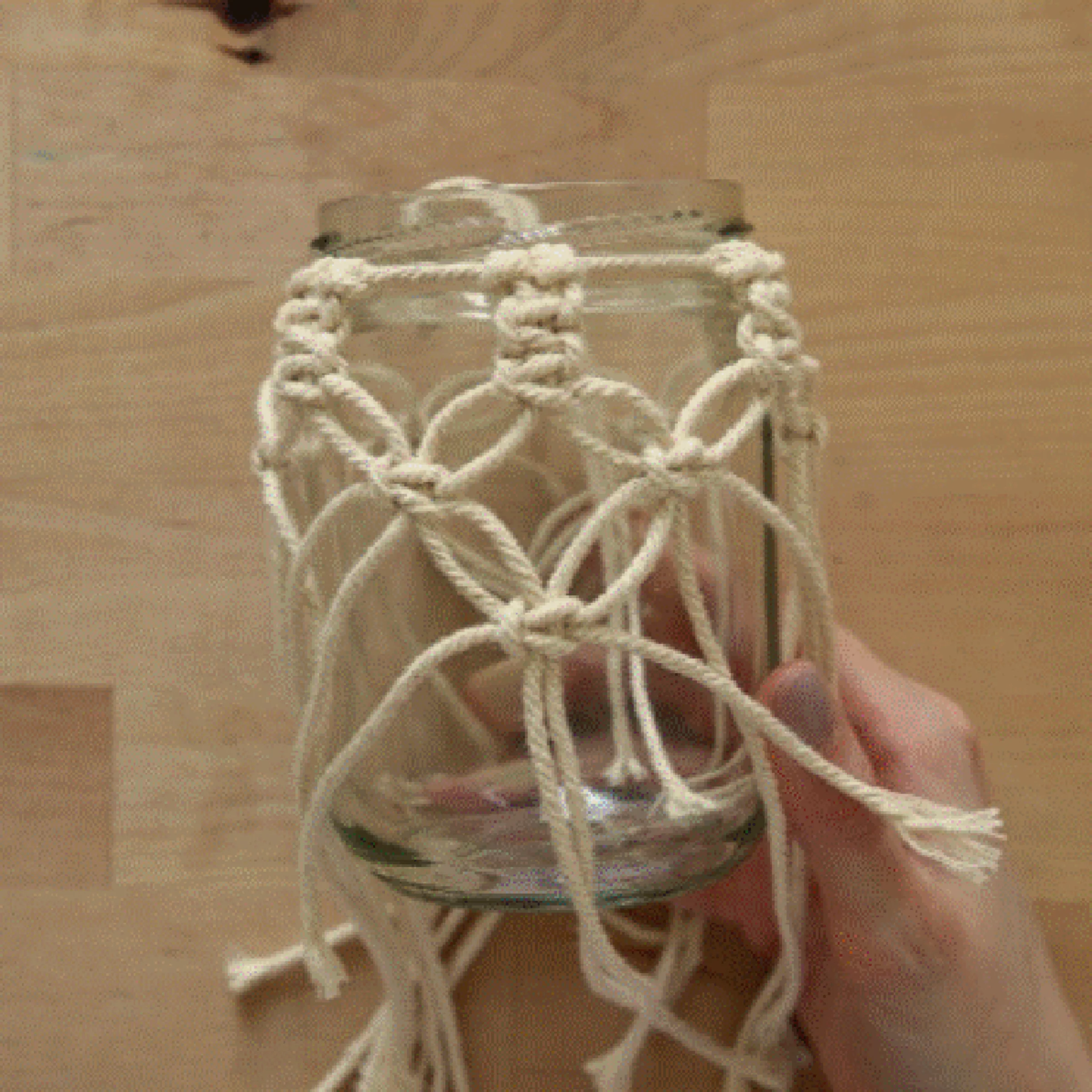Eine Hand hält ein Glas mit Makramee-Dekor auf einem Holztisch.