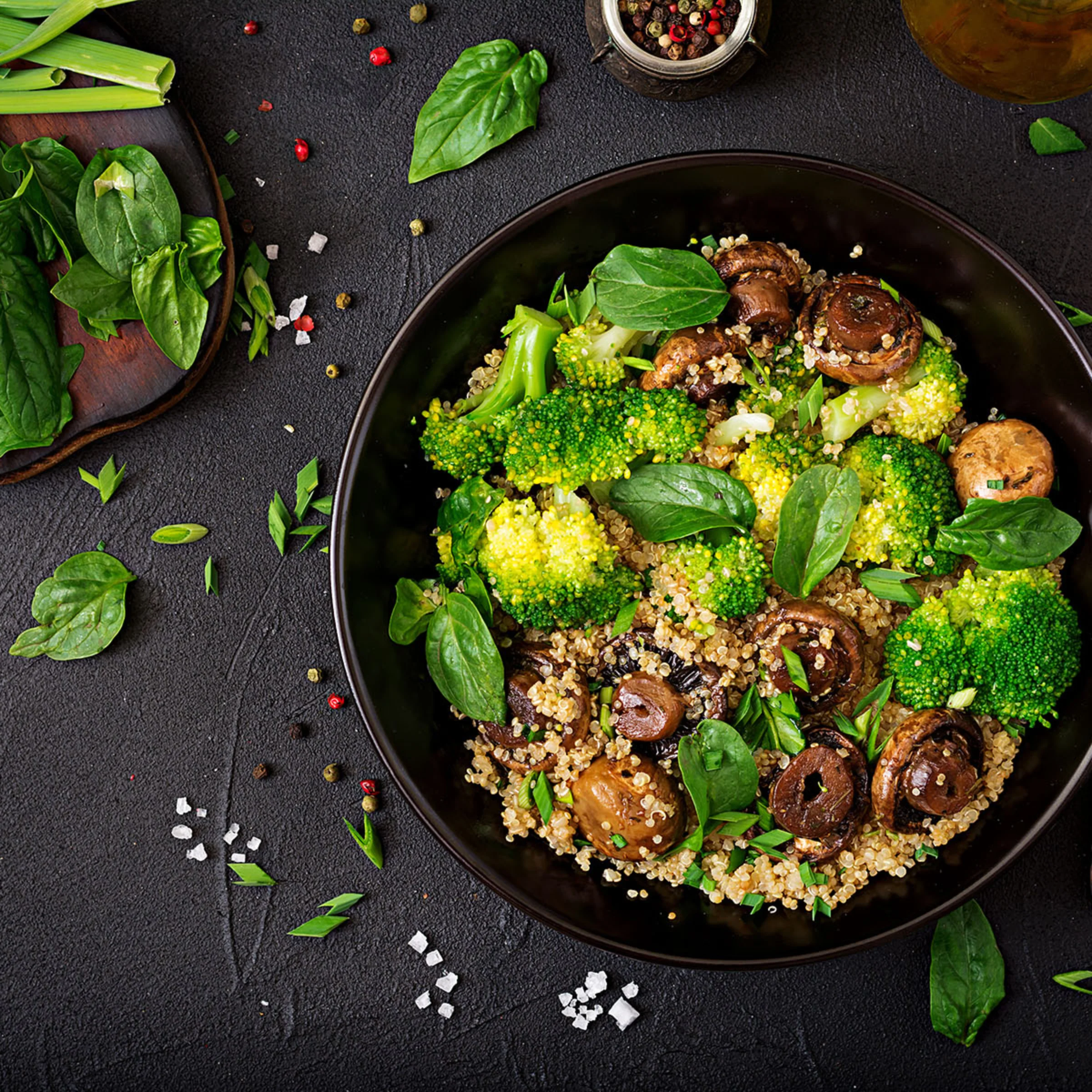 Piatto nero con insalata di quinoa, broccoli, funghi e foglie di basilico su un tavolo scuro.