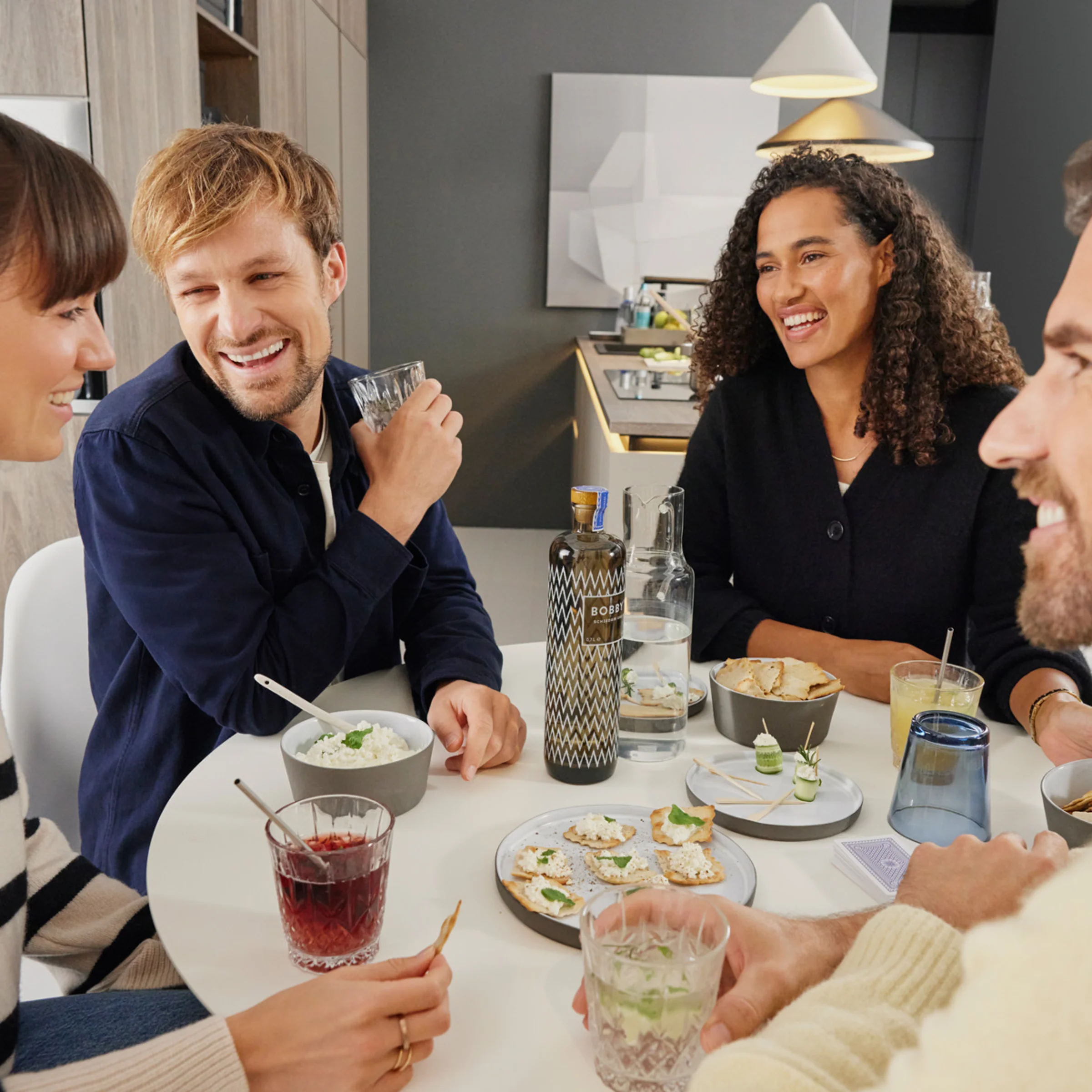 Quattro persone sedute a un tavolo con bevande e snack in un soggiorno moderno. Nolte Living.