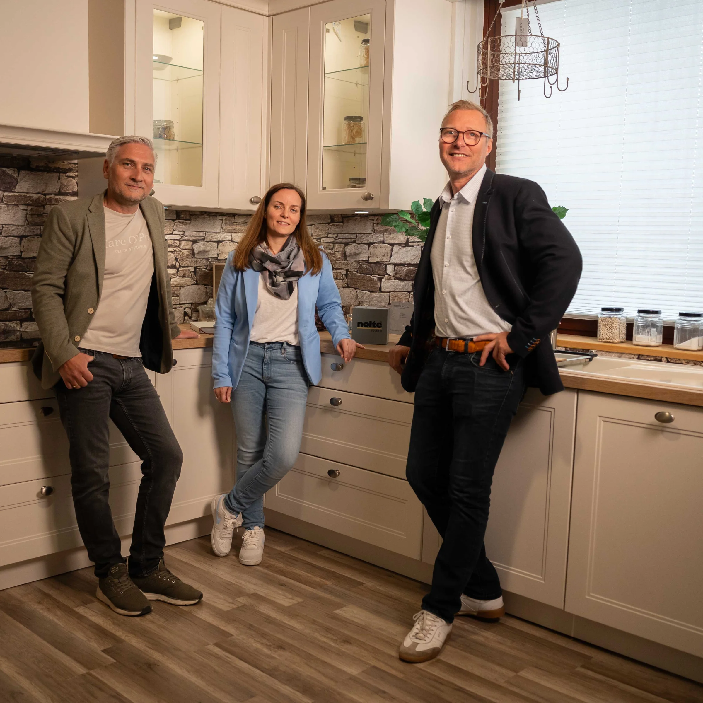 Tre persone in una cucina moderna con parete in pietra e pavimento in legno. Nolte Kitchen.