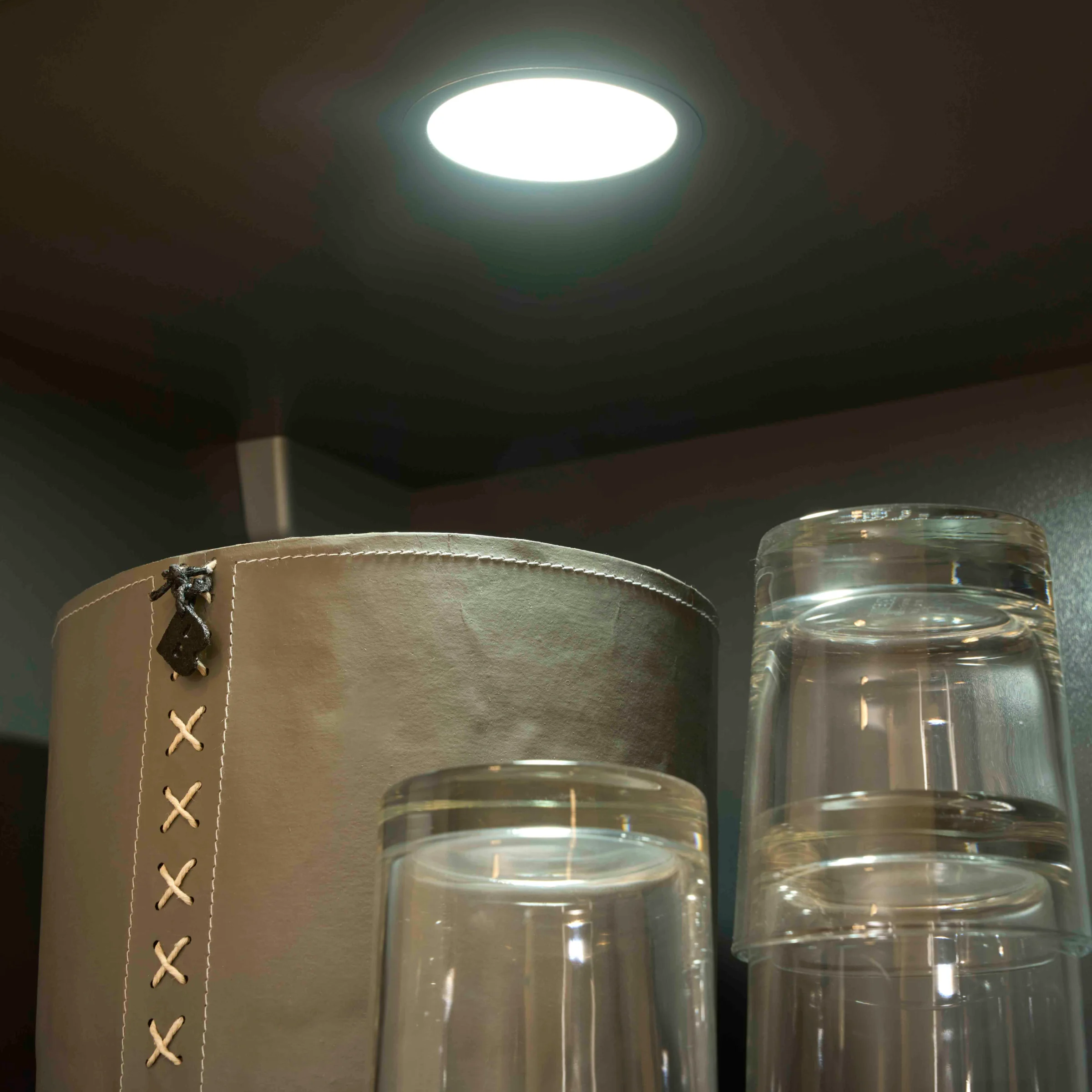 Sur une étagère, un conteneur gris et plusieurs verres sous une lampe ronde. Express Küchen.