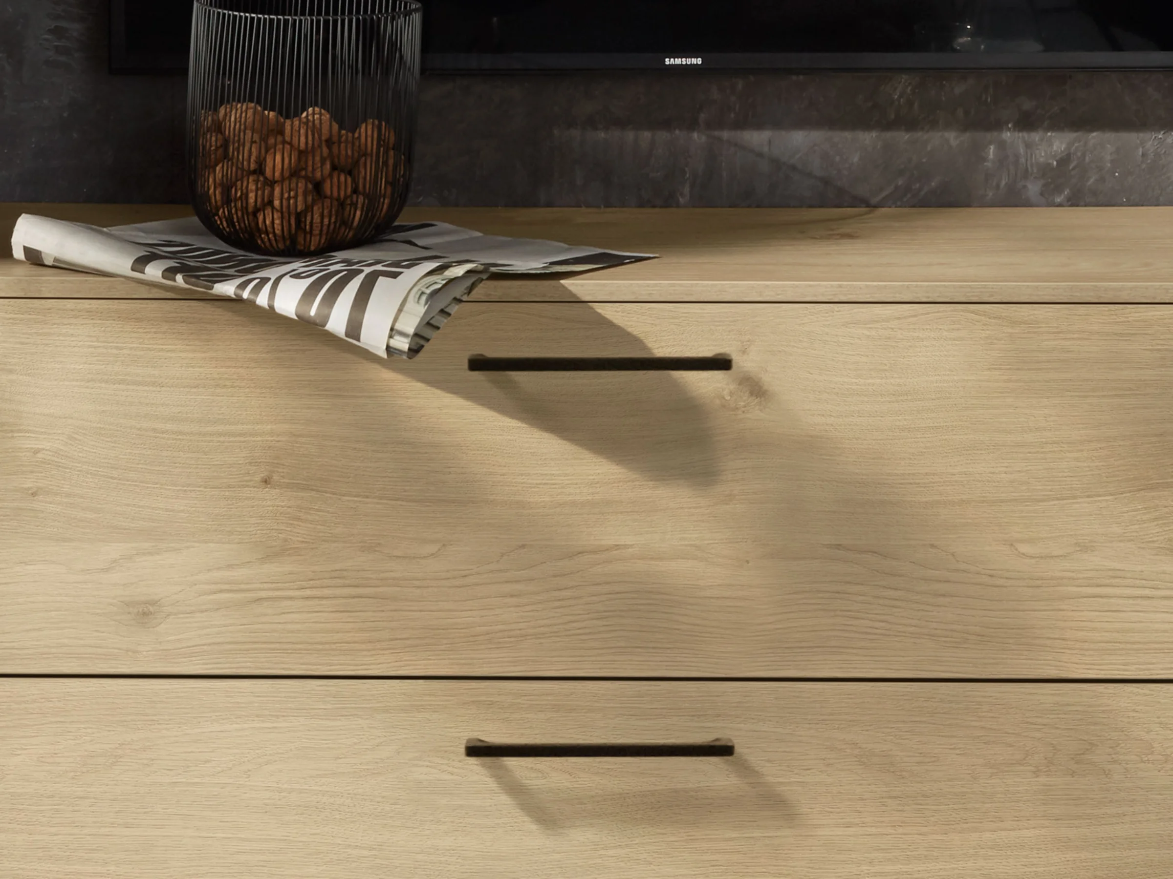 Cassettiera in legno chiaro con decorazioni nere e un giornale sopra, in stile Nolte Living.