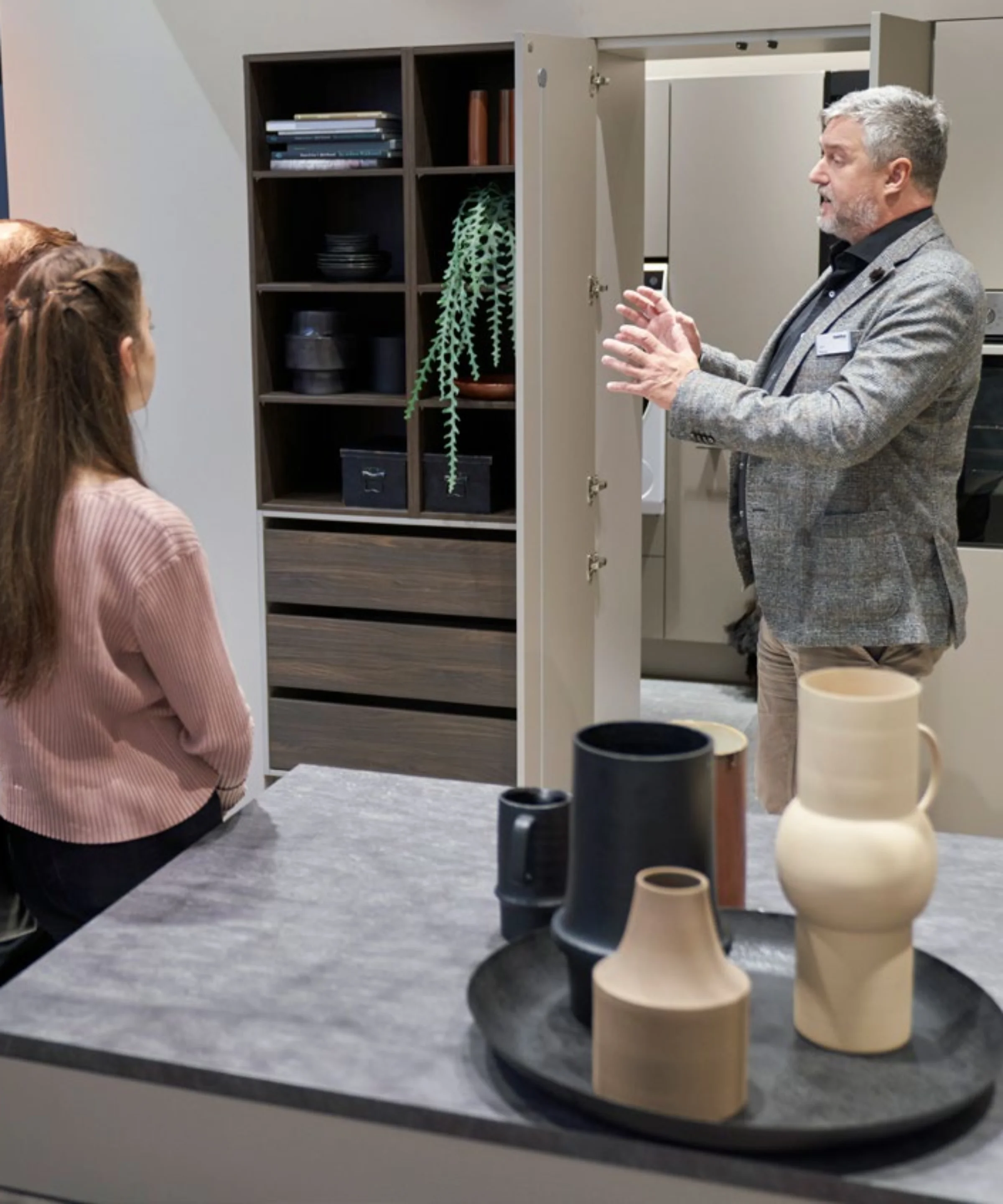 Un uomo spiega una cucina moderna a due persone, con scaffali, vasi e piante. Nolte Kitchen.