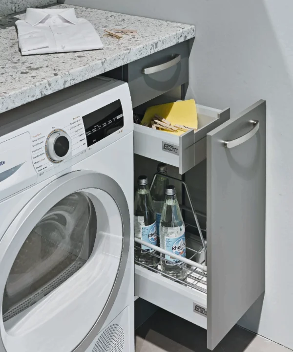 Ein Hauswirtschaftsraum mit Waschmaschine und offenem Schrank zeigt Getränke und Reinigungsmittel. Express Hauswirtschaftsräume.